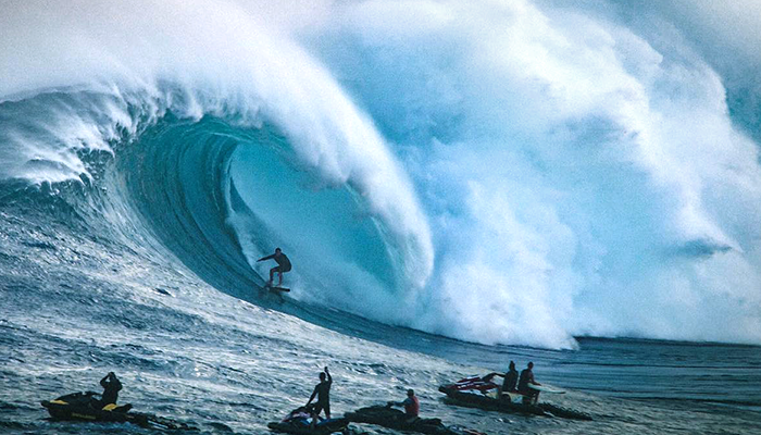 Surfer riding inside a massive ocean barrel wave while jet skis watch nearby.