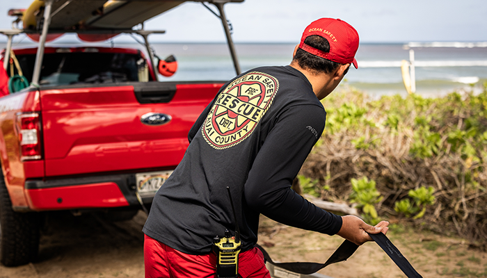 Man wearing ANETIK performance apparel preparing ocean rescue gear beside a red truck near the beach.