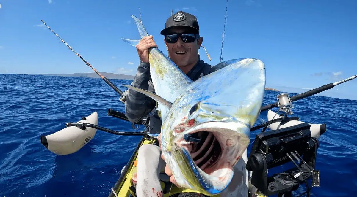 Kayak angler holding a large mahi-mahi on open blue water with fishing rods and gear visible.